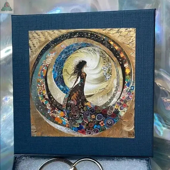 A woman in a flowing dress stands amid swirling colors, holding the Elegant Multicolor Beaded Hagstone Keychain with Goddess Box, featuring a blue, Sanibel Island–inspired frame around cosmic, fantasy-style artwork.