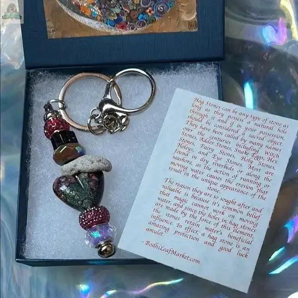 The Elegant Multicolor Beaded Hagstone Keychain with Goddess Box features a heart-shaped stone, colorful beads, and silver clasp in a blue box with a card about hag stones. An iridescent background enhances its unique appeal.