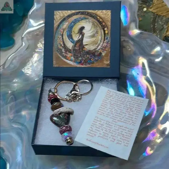 The Elegant Multicolor Beaded Hagstone Keychain with Goddess Box includes a beaded keychain, an illustrated card of a woman in a swirling dress, and a note on an iridescent surface, capturing the dreamy spirit of Sanibel Island.