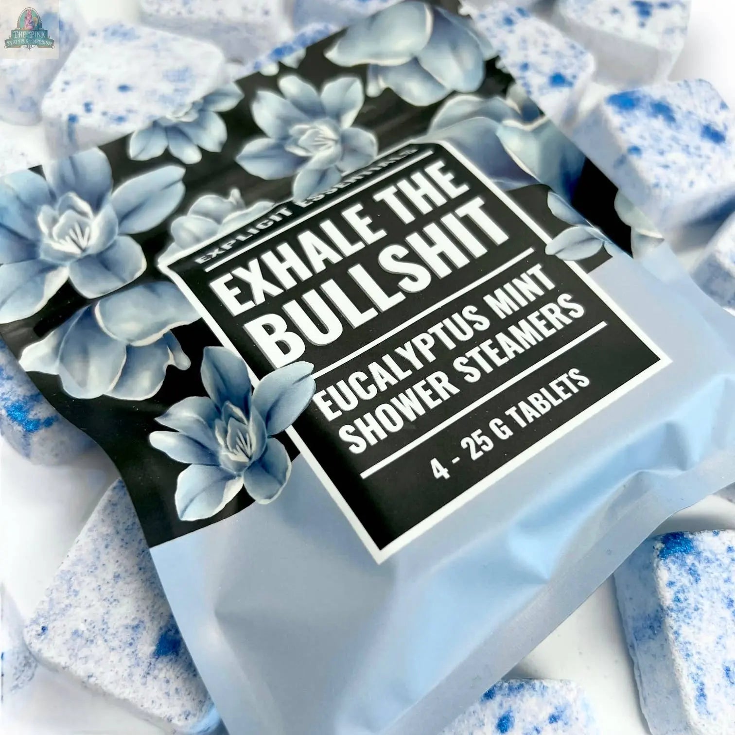 A package of Explicit Essentials’ Exhale The Bullsh!t Shower Steamers with blue floral designs and bold white lettering rests on a surface scattered with blue and white speckled tablets.