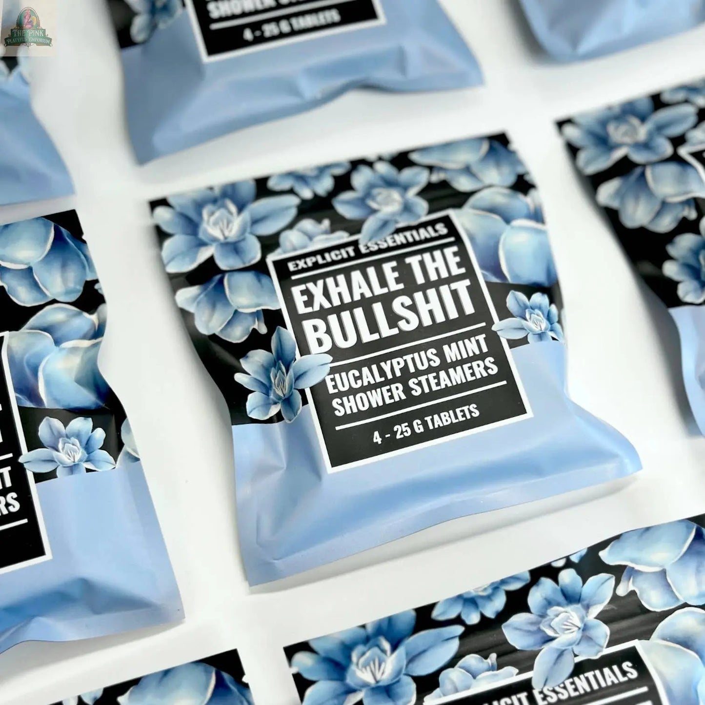 A close-up shows light blue Exhale The Bullsh!t Shower Steamers by Explicit Essentials, each package decorated with blue flowers and labeled Eucalyptus Mint. Four tablets per pack are visible.