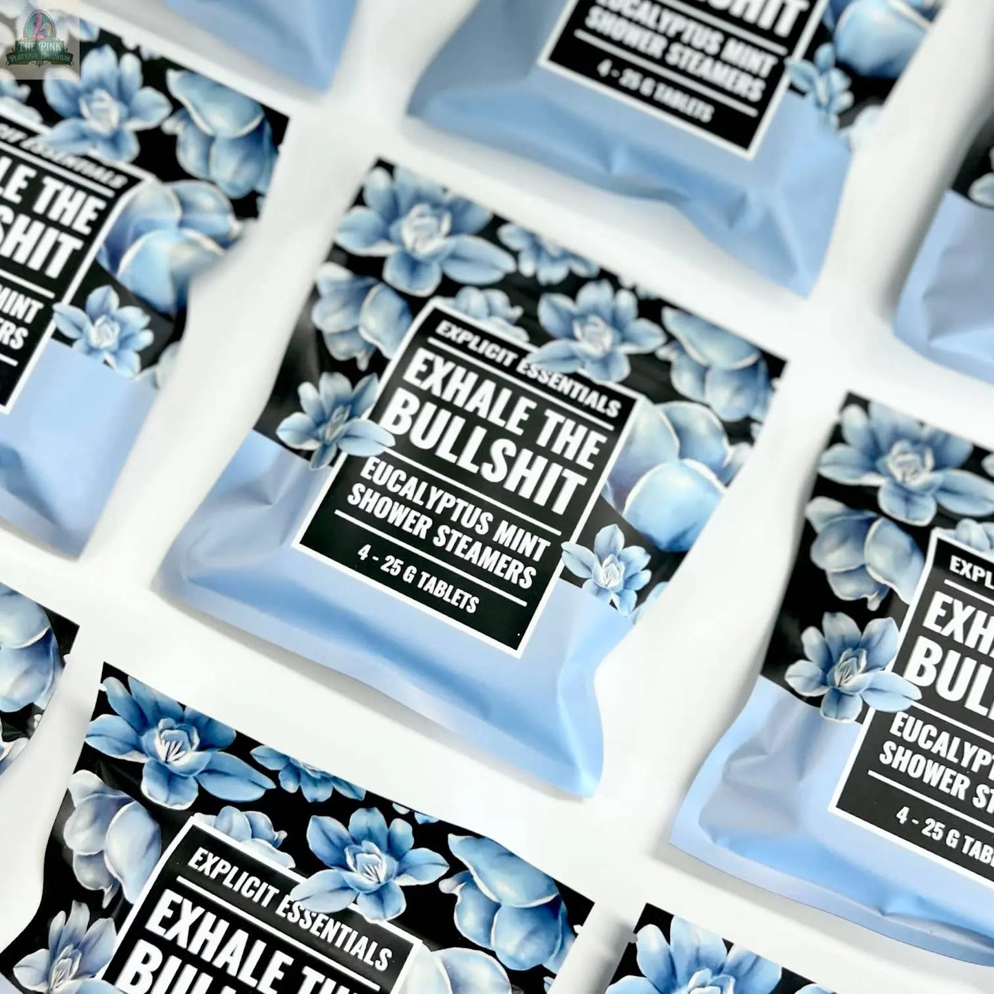 Several blue and black packets of Explicit Essentials' Exhale The Bullsh!t Shower Steamers are arranged in a grid. The all-natural packaging features blue flowers and bold white text on a black background.
