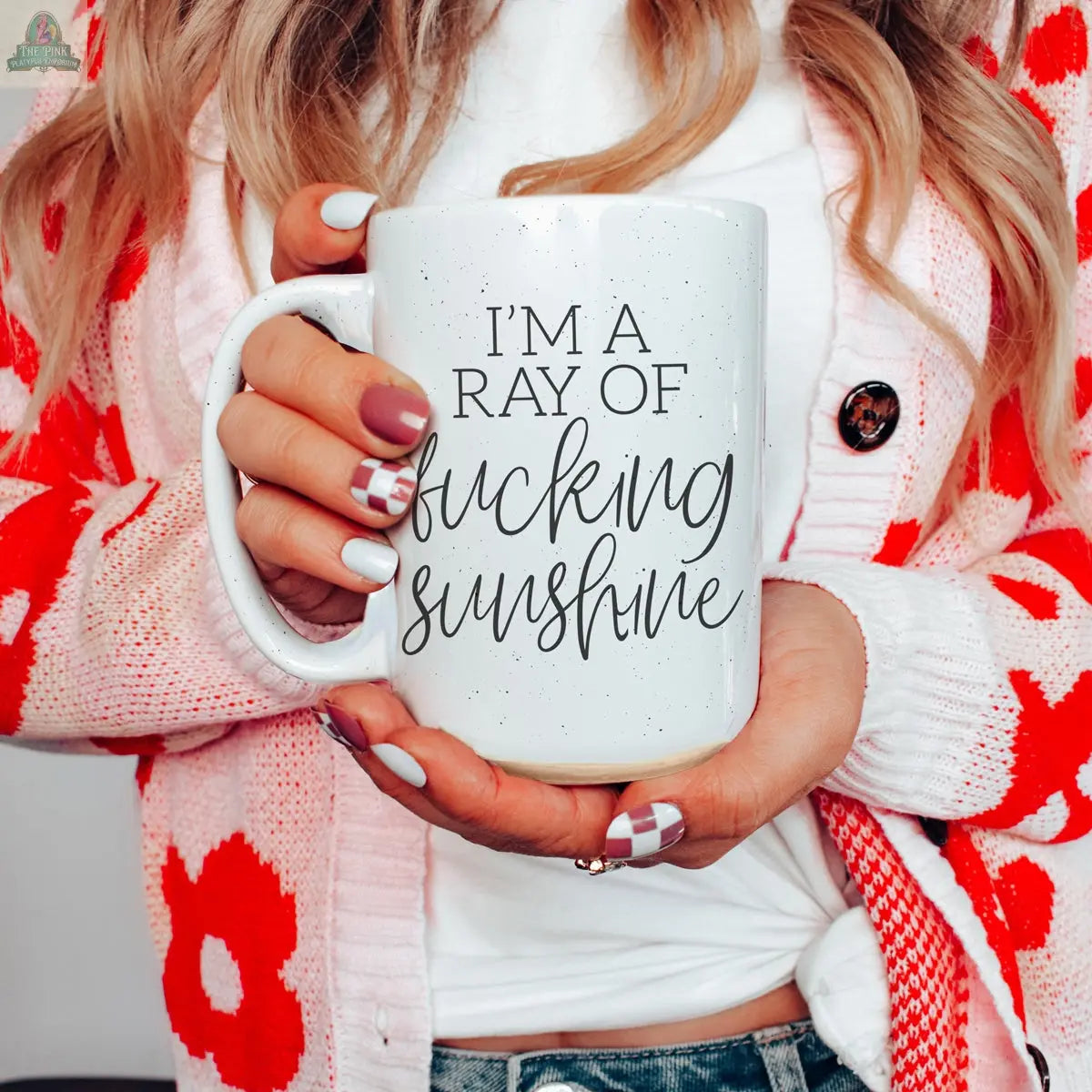 A woman with long hair holds the F'n Sunshine 16oz ceramic mug that says "IM A RAY OF fucking sunshine." She wears a white top, pink cardigan with red flowers, and has white and pink painted nails.