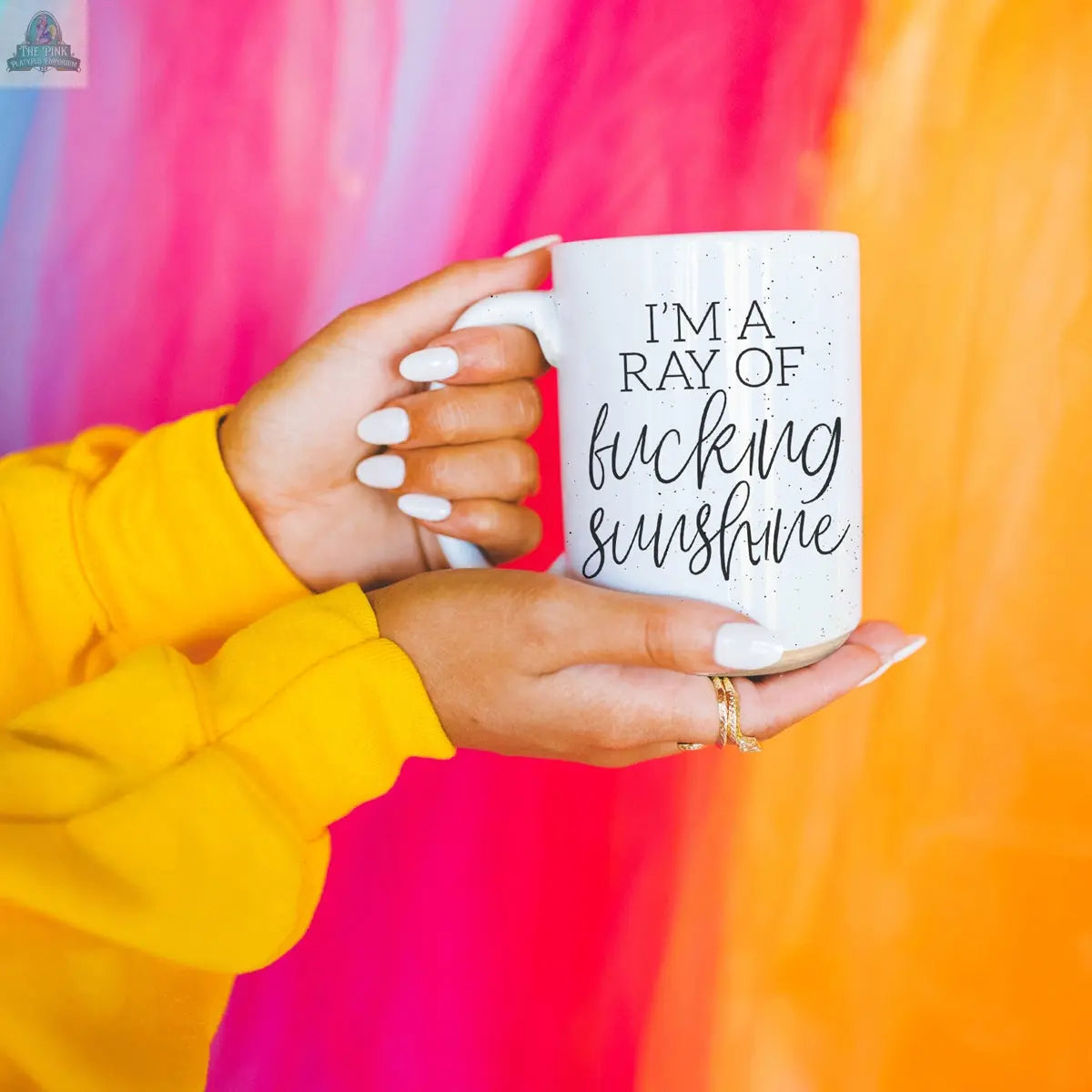 Someone in a yellow sweater holds the F'n Sunshine 16oz ceramic mug, featuring bold black text, against a vibrant, blurred background of pink, red, orange, and blue.