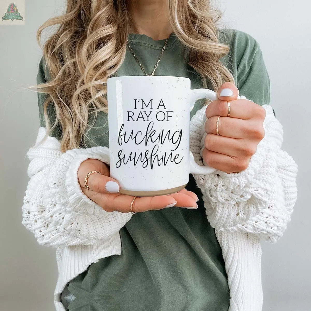 A woman with long wavy hair holds the F'n Sunshine 16oz ceramic mug that says "IM A RAY OF f***ing sunshine." She wears a green shirt, a chunky white sweater, and several rings.