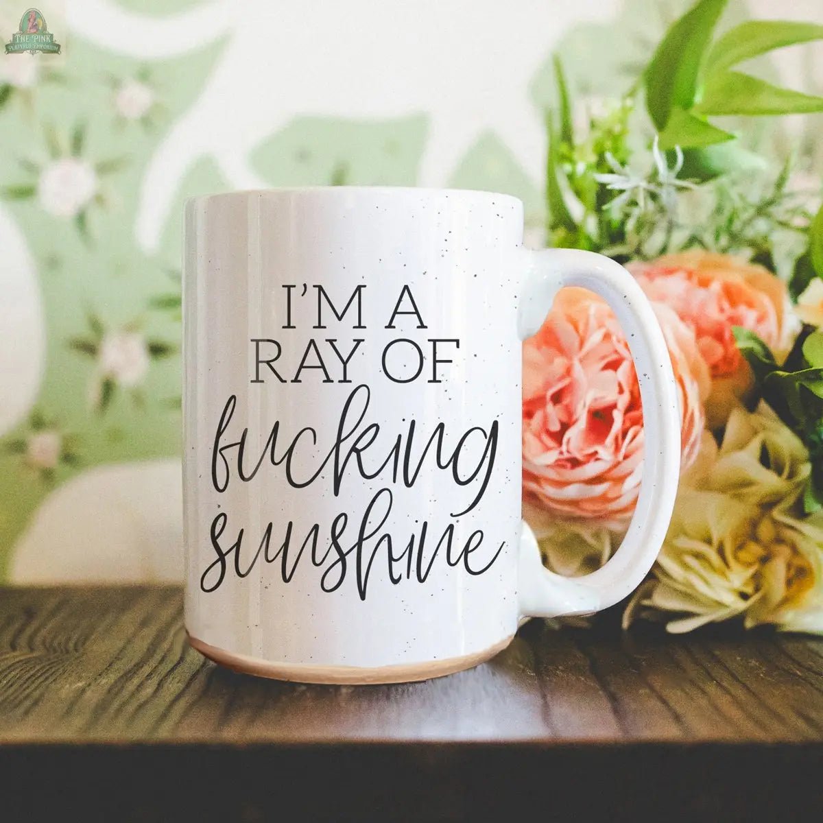 The F'n Sunshine 16oz white ceramic mug with black text sits on a wooden surface beside a bouquet of orange and cream flowers, with green leaves in the background.