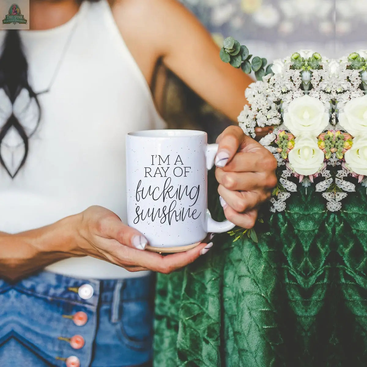 A person in a white sleeveless top and blue jeans holds the F'n Sunshine 16oz ceramic mug, featuring the message "IM A RAY OF fucking sunshine," next to a bouquet of white roses and greenery.