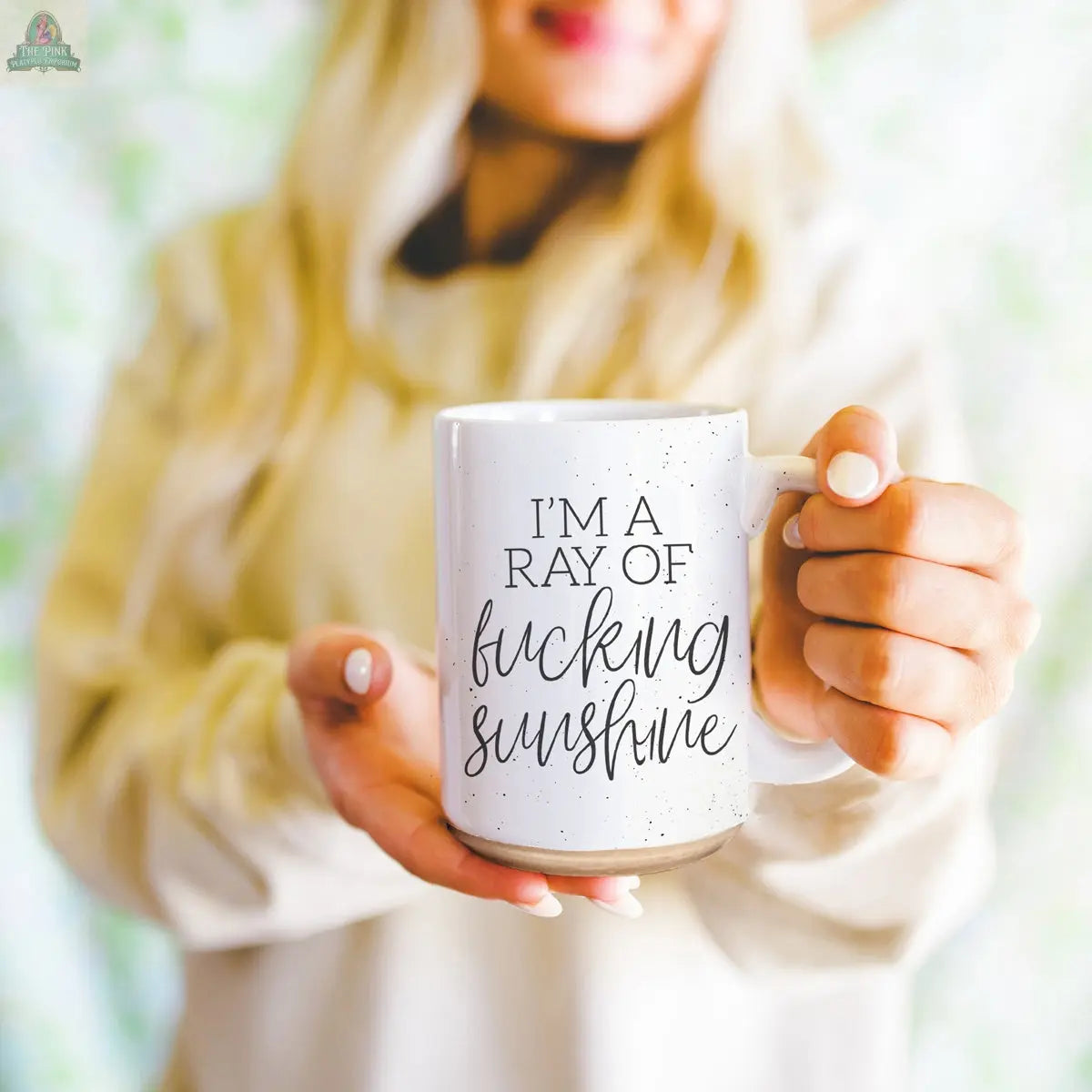 A woman with long blonde hair holds the F'n Sunshine 16oz ceramic mug featuring the quote, “I’m a ray of fucking sunshine,” with softly blurred green foliage in the background.