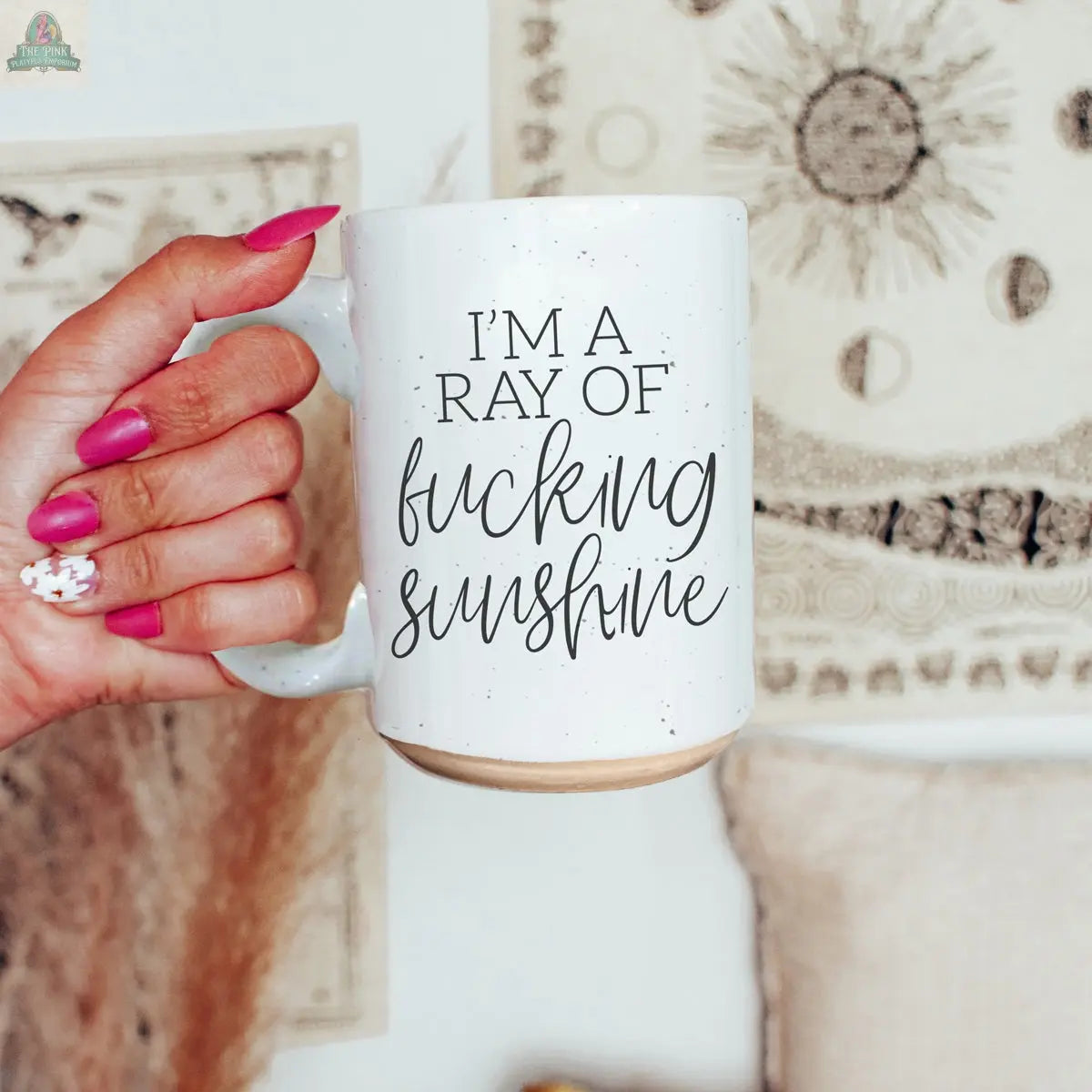 A hand with pink and white painted nails holds the F'n Sunshine 16oz ceramic mug featuring bold black text. Sun and geometric design tapestries hang in the background.