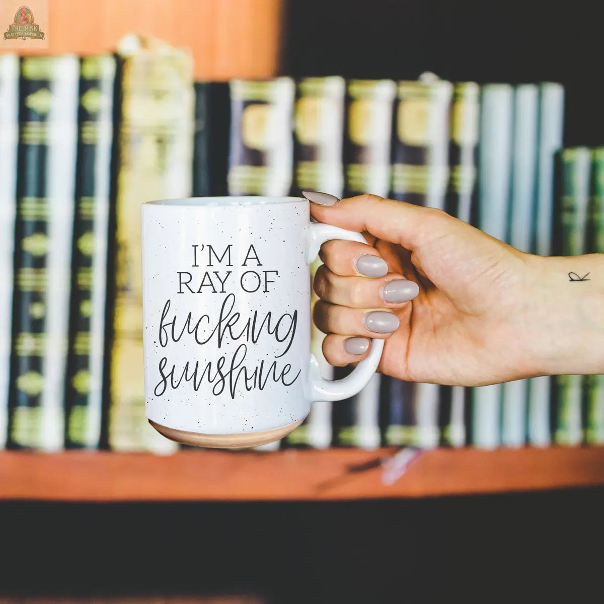 A hand with light polish holds the F'n Sunshine 16oz mug featuring bold "IM A RAY OF fucking sunshine" text, against a blurred bookshelf background.