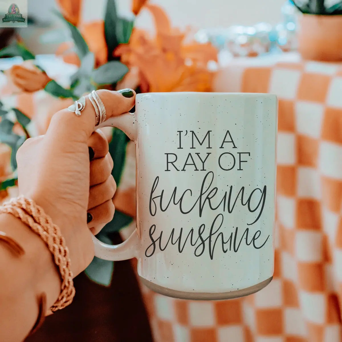 A hand holds the F'n Sunshine 16oz mug with "IM A RAY OF fucking sunshine" in bold black letters. Vibrant orange flowers and an orange and white checkered tablecloth add a cheerful background.