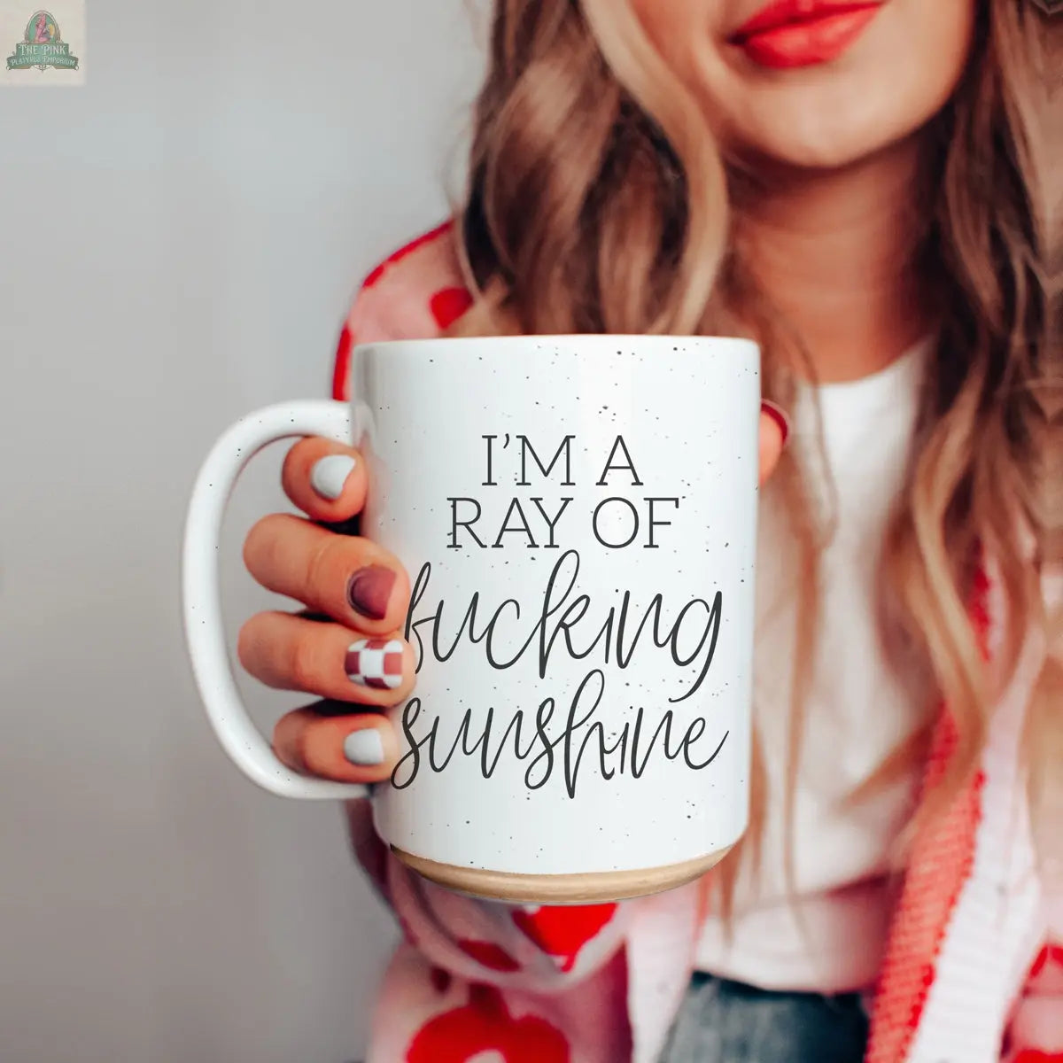 A woman with long hair and red lipstick holds the F'n Sunshine 16oz mug, which reads, "I'M A RAY OF FUCKING SUNSHINE." Sporting painted nails and a colorful sweater, she radiates positivity and confident style.