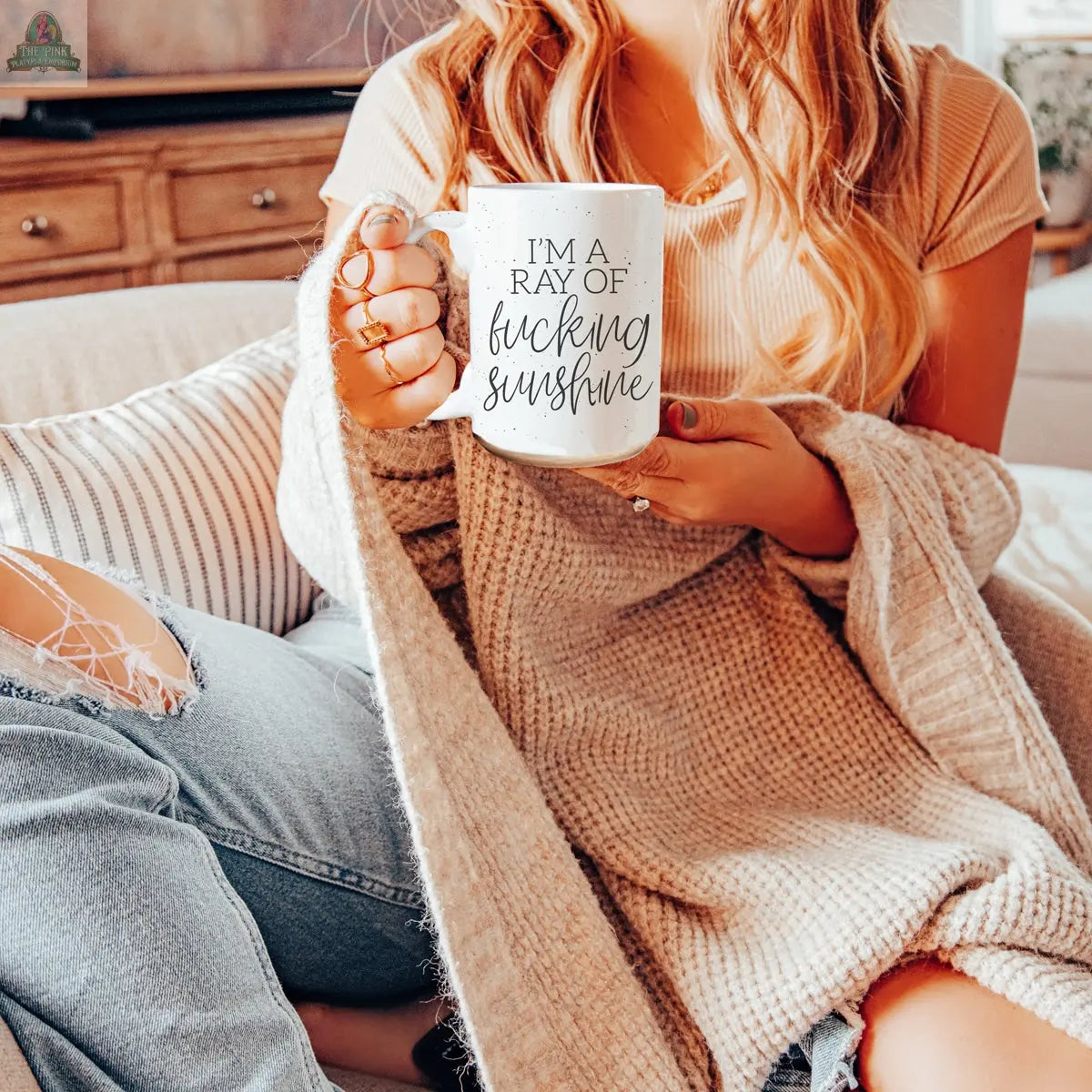 A woman with long, wavy hair sits casually on a couch in a T-shirt and ripped jeans, wrapped in a beige blanket and holding the F'n Sunshine 16oz mug that reads, "IM A RAY OF f*cking SUNSHINE.