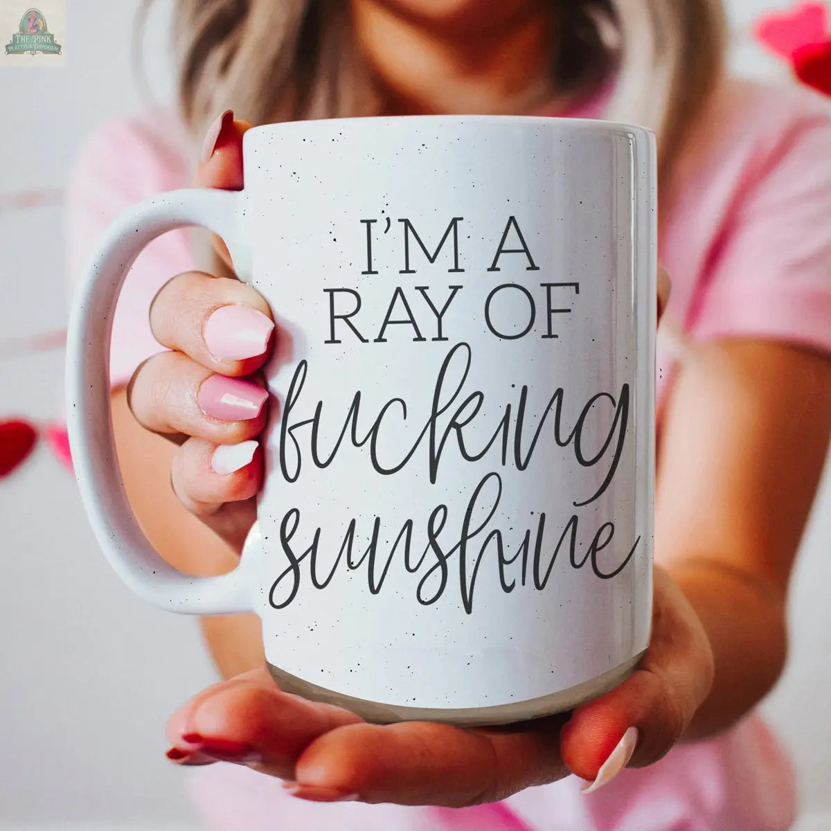 Wearing a pink shirt, a person holds the F'n Sunshine 16oz ceramic mug featuring bold black text, "I'M A RAY OF f*cking SUNSHINE," standing before a softly blurred background with red and pink hues.