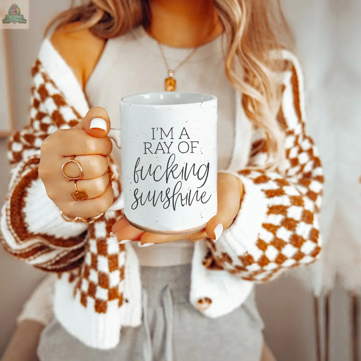 A woman holds the F'n Sunshine 16oz ceramic mug, featuring bold black text reading "I'M A RAY OF FUCKING SUNSHINE," while wearing a brown and white checkered cardigan. Only her torso and hands are shown.