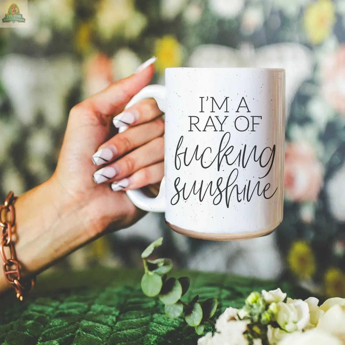 A hand with manicured nails holds the F'n Sunshine 16oz ceramic mug, featuring bold black text, "IM A RAY OF fucking sunshine," against a backdrop of blurred floral and green accents.