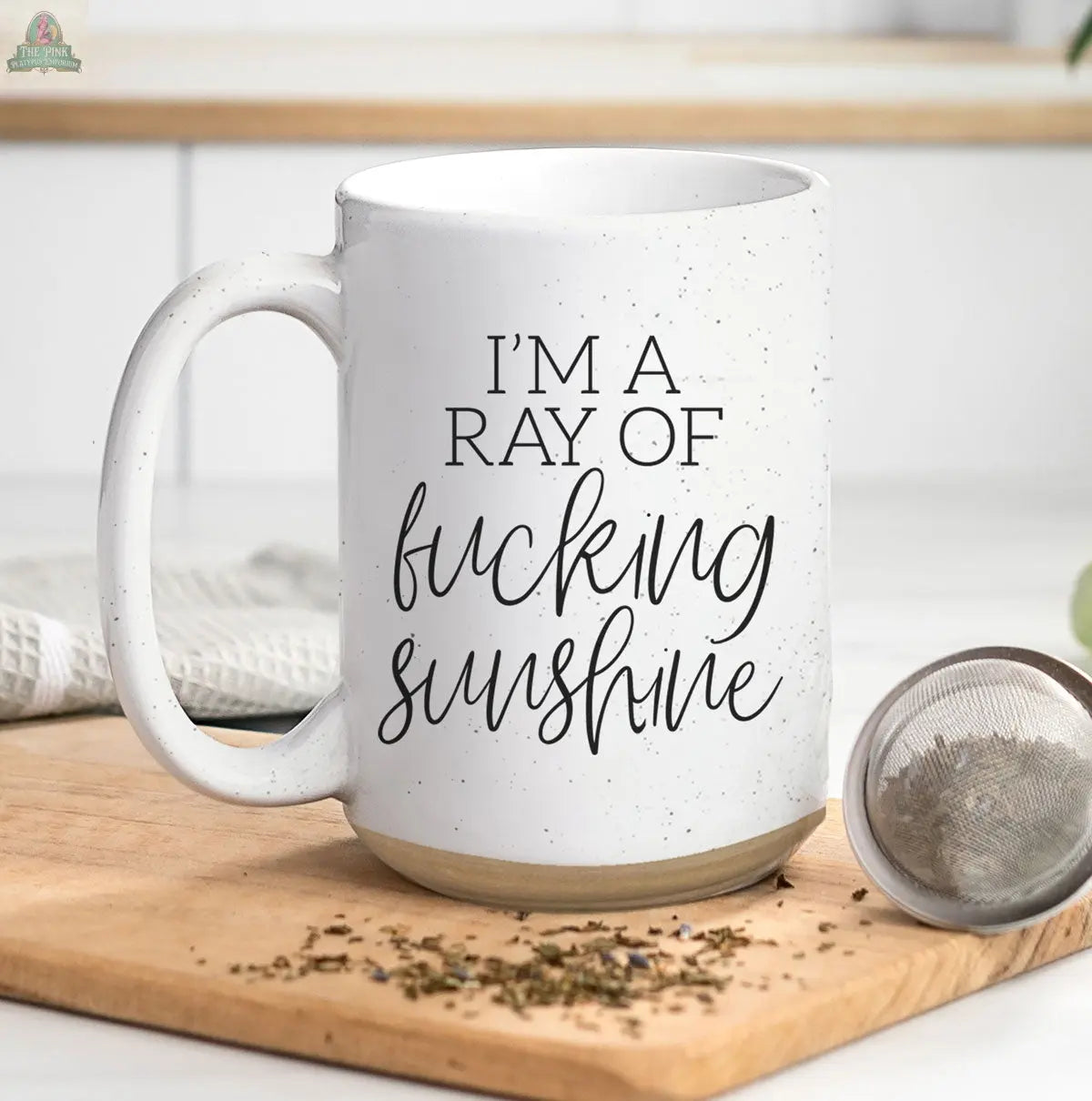 The F'n Sunshine 16oz white ceramic mug sits on a wooden board, showing “I’m a ray of fucking sunshine” in black script. A tea infuser and a white kitchen towel lie nearby on a light-colored countertop.