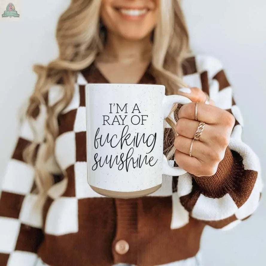 A smiling woman with long, wavy hair holds the F'n Sunshine 16oz ceramic mug that reads "I’m a ray of fucking sunshine." She wears a brown and white checkered cardigan and gold rings.