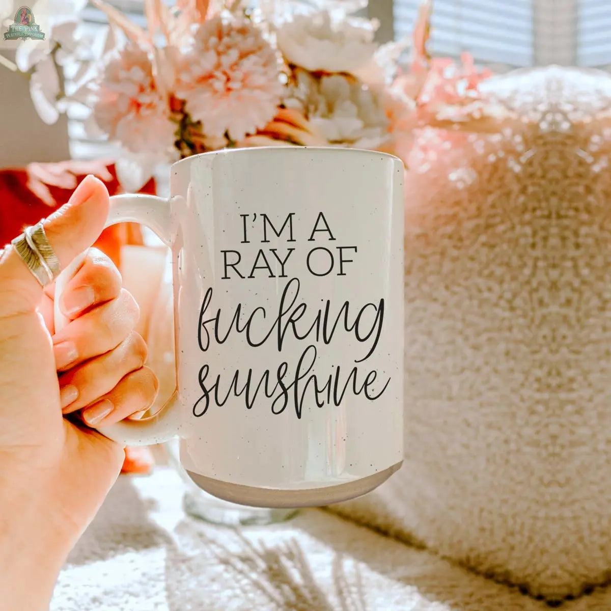A hand holds the F'n Sunshine 16oz ceramic mug with bold "IM A RAY OF fucking sunshine" text. Soft light highlights pink flowers and a textured beige pillow in the background.