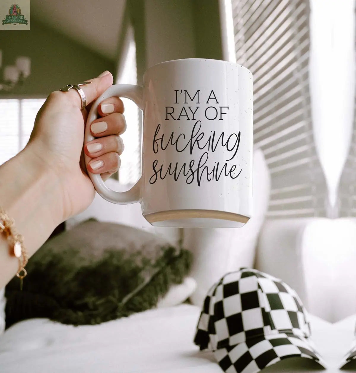 A hand holds the "F'n Sunshine 16oz" inspirational ceramic mug; a checkered cap and soft cushions rest on a bed in the softly lit background.