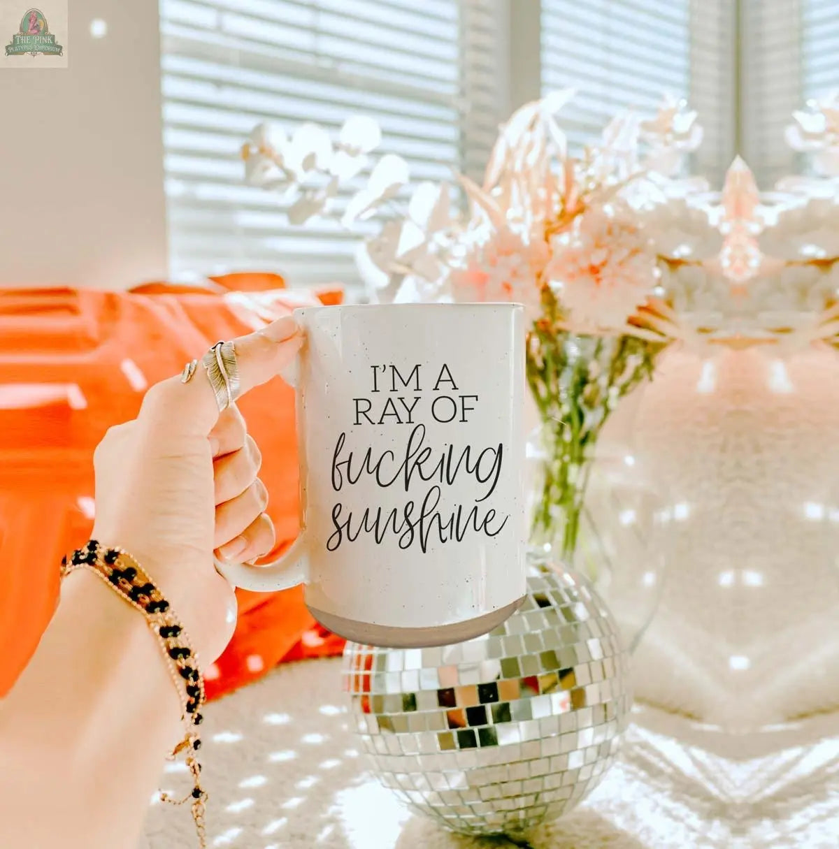 A hand holds the F'n Sunshine 16oz ceramic mug with bold text, set against a sunlit room featuring white flowers in a vase and a disco ball on a textured surface.