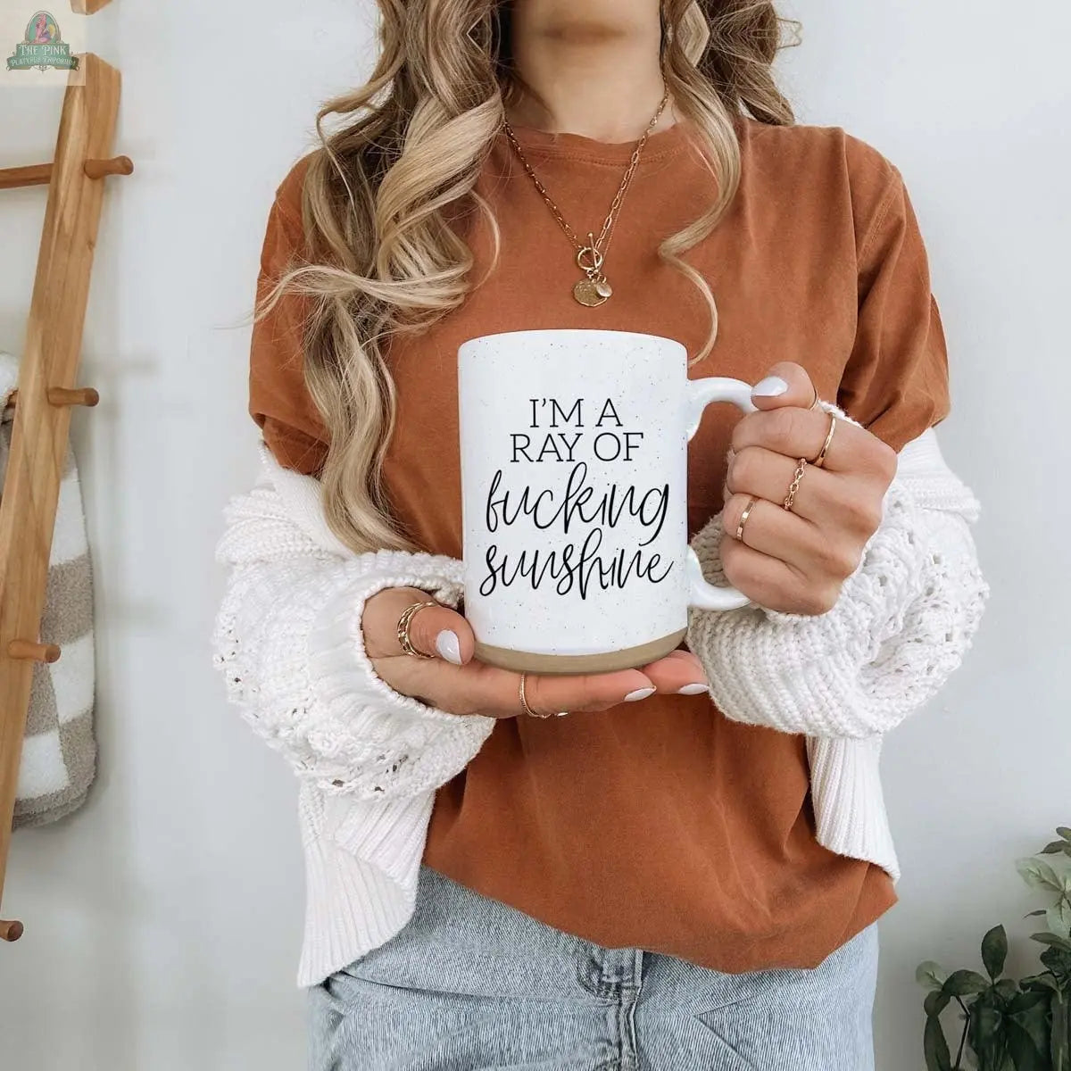 A woman with wavy hair, wearing a brown shirt, white cardigan, and jeans, stands indoors by a wooden ladder holding the F'n Sunshine 16oz inspirational ceramic mug.