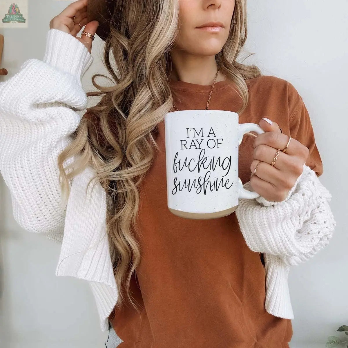 A woman with long wavy hair holds the F'n Sunshine 16oz inspirational ceramic mug that reads "IM A RAY OF fucking SUNSHINE." She wears a brown shirt, white sweater, rings, and a hat that partially covers her face.