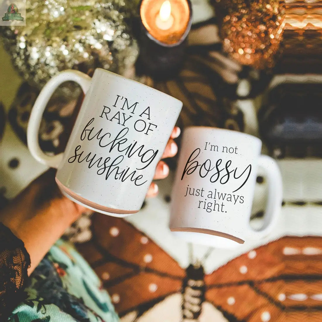 Two 16oz F'n Sunshine mugs—one saying "IM A RAY OF fucking sunshine," the other "Im not bossy just always right"—are held above a decorative surface with a candle and festive decor in the background.