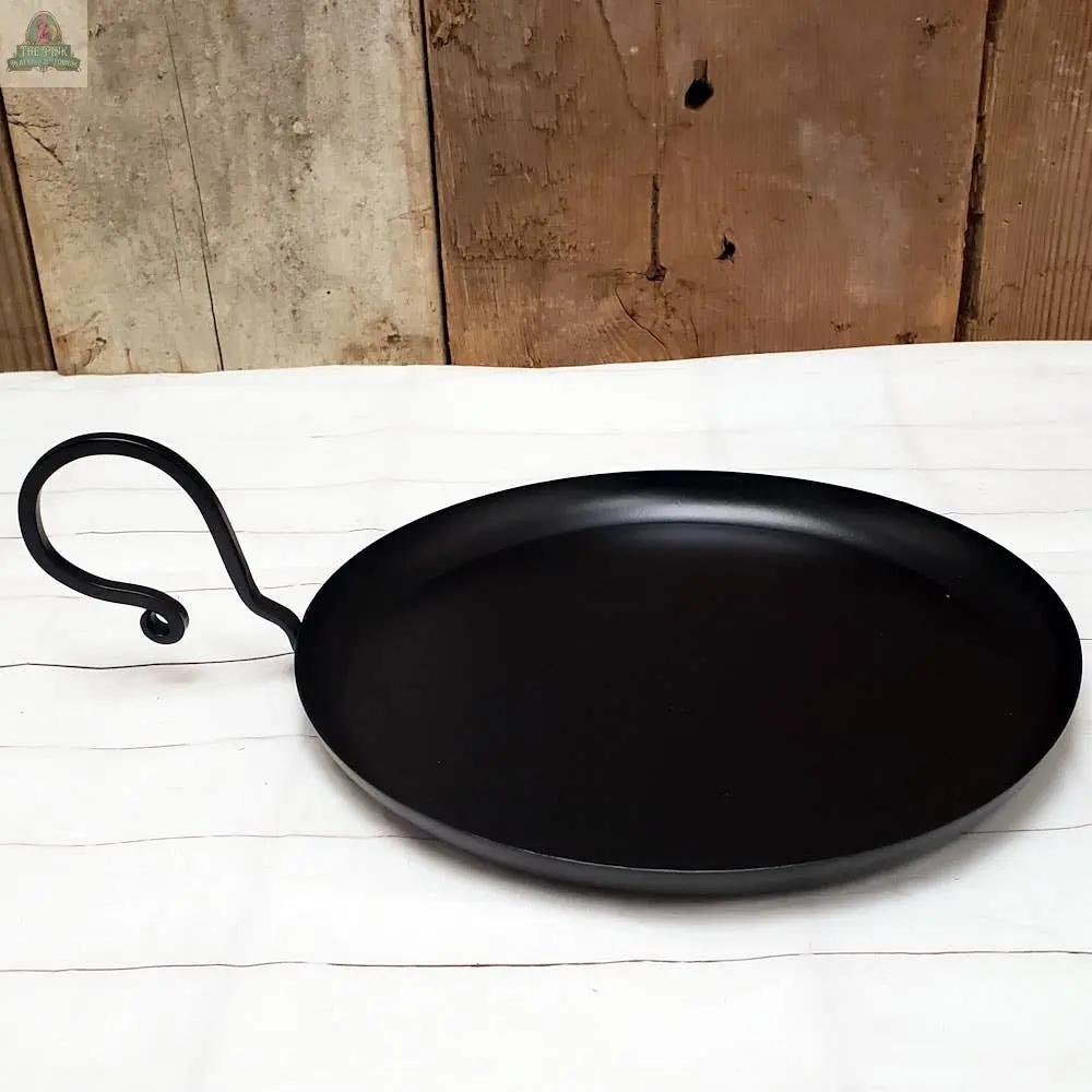 The Flat Metal Pan With Handle, featuring a round black iron surface and a curved handle, rests on a white cloth and stands out against a rustic wooden background.