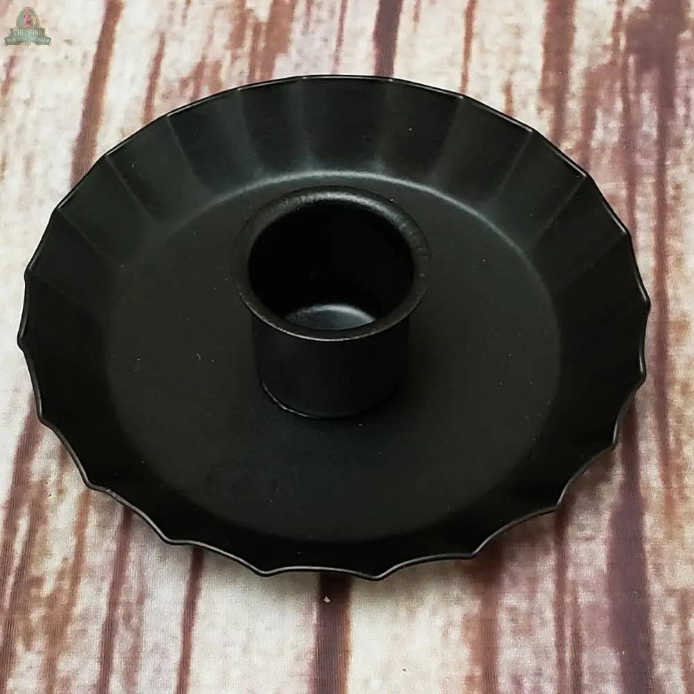 The Fluted Edge Taper Holder: Black features a black metal design with a fluted edge and central cylindrical section, perfect for displaying a single taper candle. It sits elegantly on a striped wooden surface.