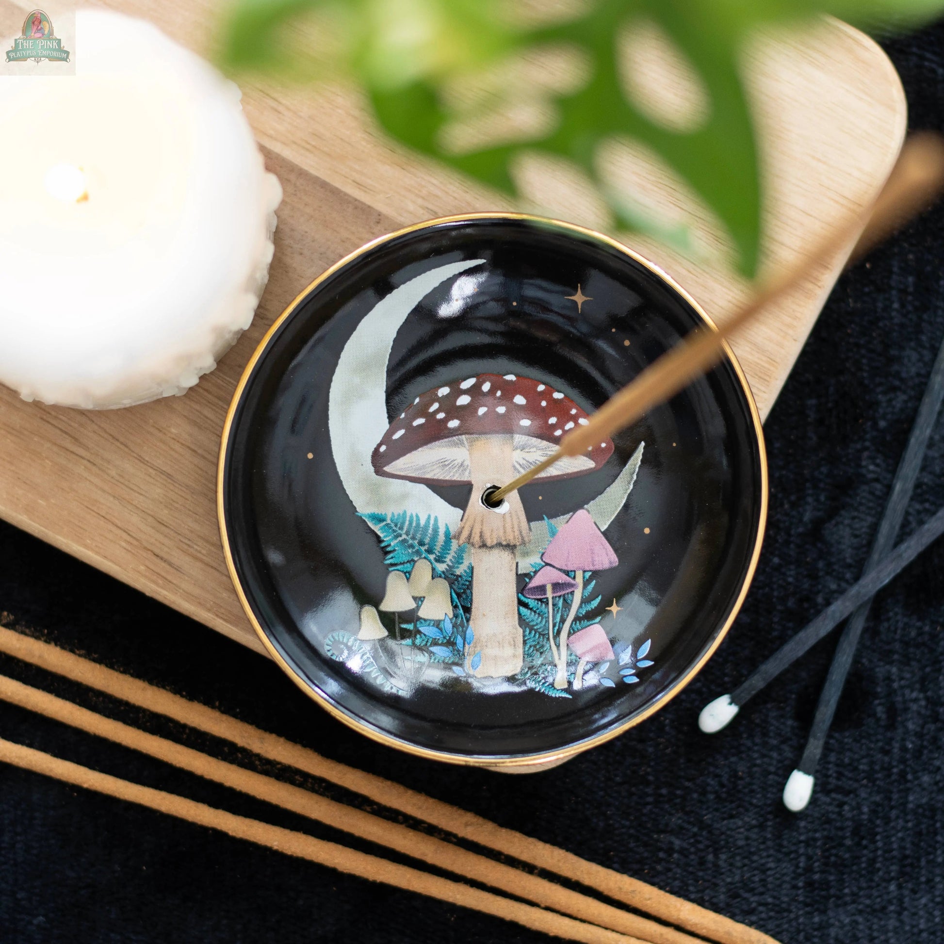 The Forest Mushroom Ceramic Incense Holder Plate, featuring moon and mushroom artwork, holds a burning incense stick beside a lit candle on wood with green leaves—ideal for mystical woodland gifts and unique home decor.