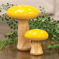 Two yellow mushroom-shaped decor pieces with wooden bases on a wooden surface with greenery in the background.