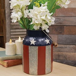 Decorative patriotic milk can vase with American flag design on a wooden surface with candles and flowers.