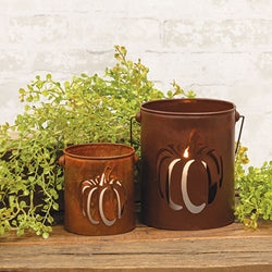 Two rust-colored metal lanterns with pumpkin designs on a wooden surface with greenery.