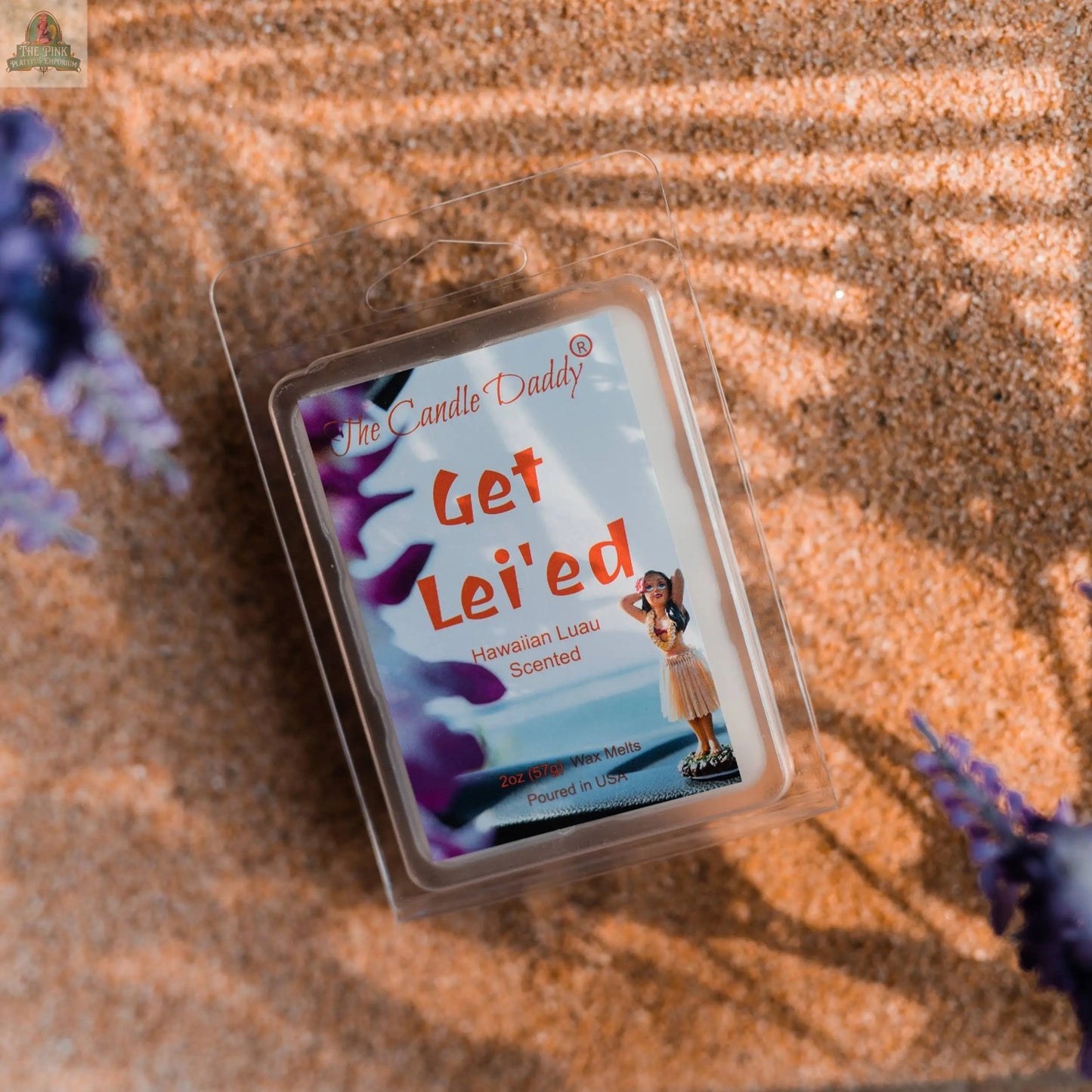 A pack of GET LEI'ED - HAWAIIAN LUAU SCENTED WAX MELT (2oz) rests on sand with purple flowers and sunlight, featuring packaging with a hula dancer—ideal for aromatherapy wax melt lovers.