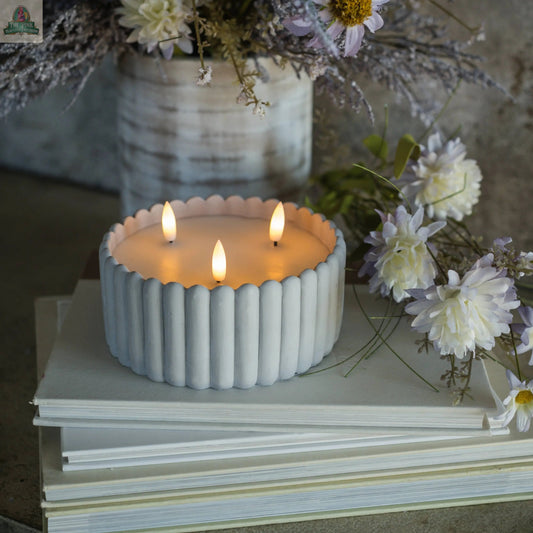 A GREY CEMENT 3 WICK 3D FLAME CANDLE burns atop a stack of books, with light purple and white flowers in the background and beside the candle.