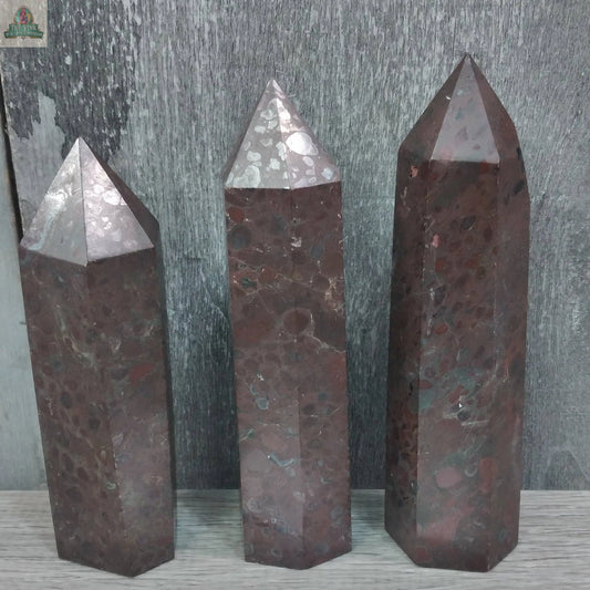 Three upright Keystone Crystals Gemstone Obelisks – 4 to 5 oz Bulk Crystal Towers in Coffee Bean Jasper, featuring polished, dark reddish-brown hexagonal shapes with speckled patterns, are shown against a gray wooden background.