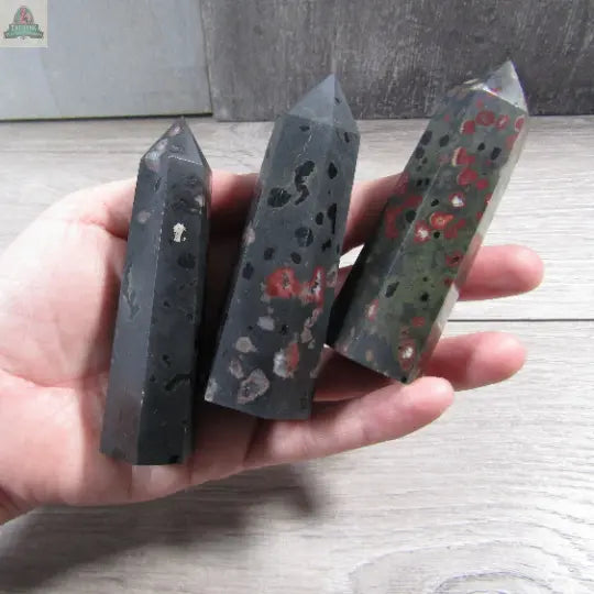 A hand holds three polished Coffee Bean Jasper Gemstone Obelisks by Keystone Crystals (4 to 5 oz bulk crystal towers), featuring dark bases with red speckles, against a light wooden background.