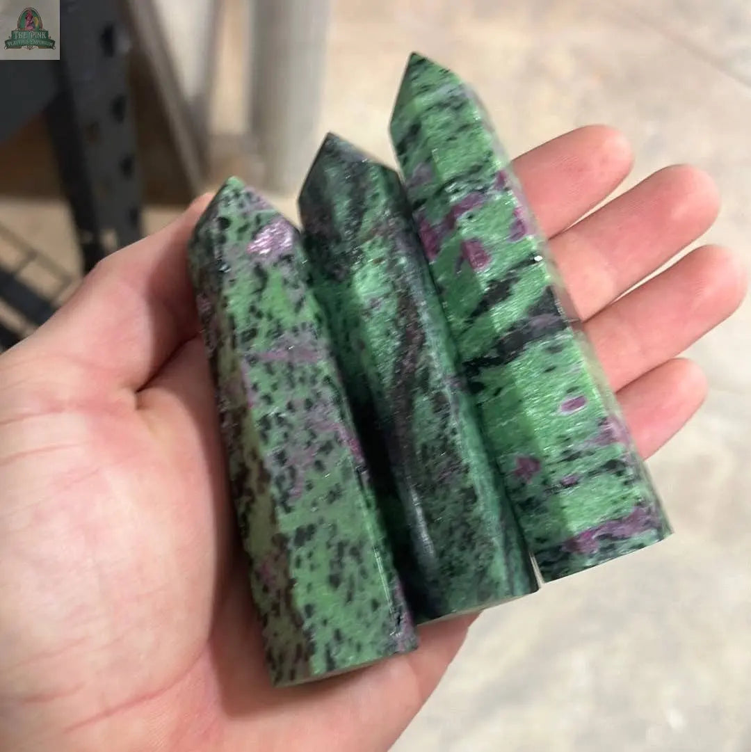 A hand holds three Keystone Crystals Coffee Bean Jasper obelisks (4–5 oz), featuring gemstone towers that showcase a beautiful mix of mottled green, black, and purple-pink patterns.