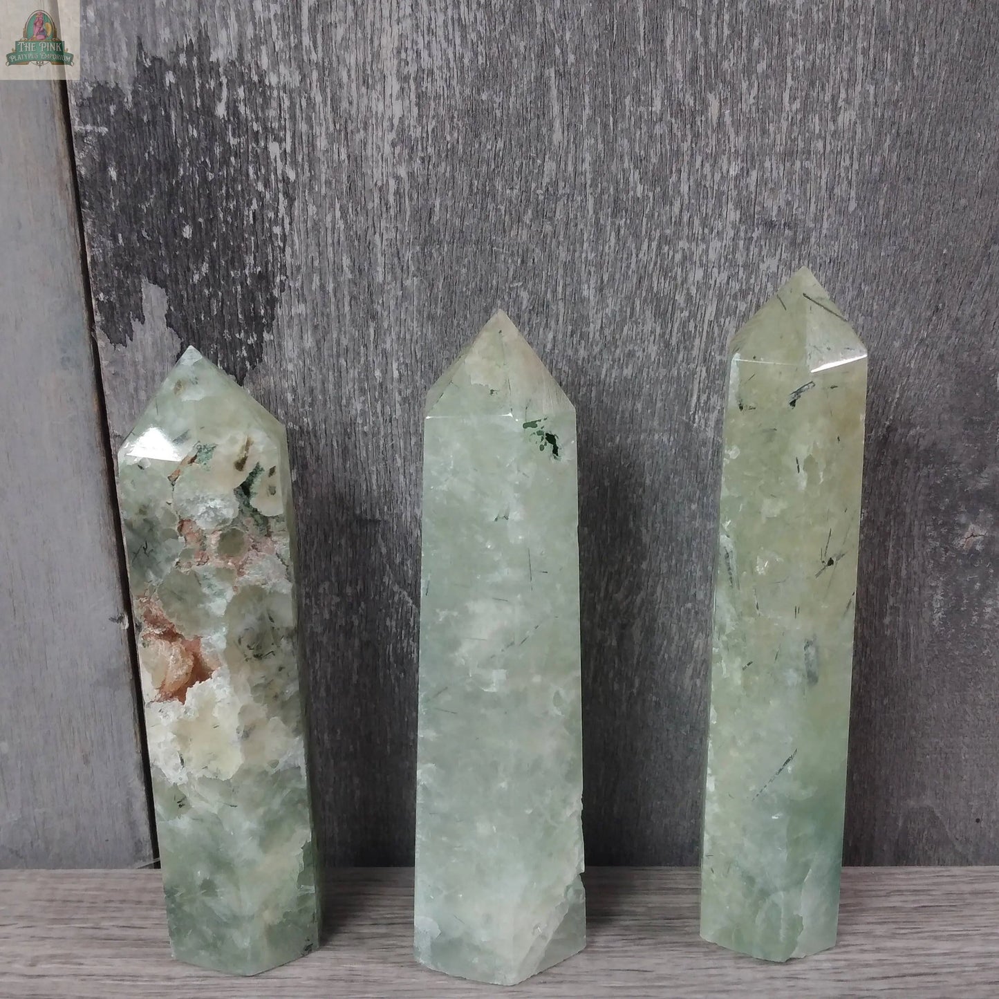 Three Coffee Bean Jasper obelisks from Keystone Crystals stand upright on a gray wooden surface with a textured gray wood background. These bulk 4 to 5 oz gemstone towers are part of the Gemstone Obelisks collection.