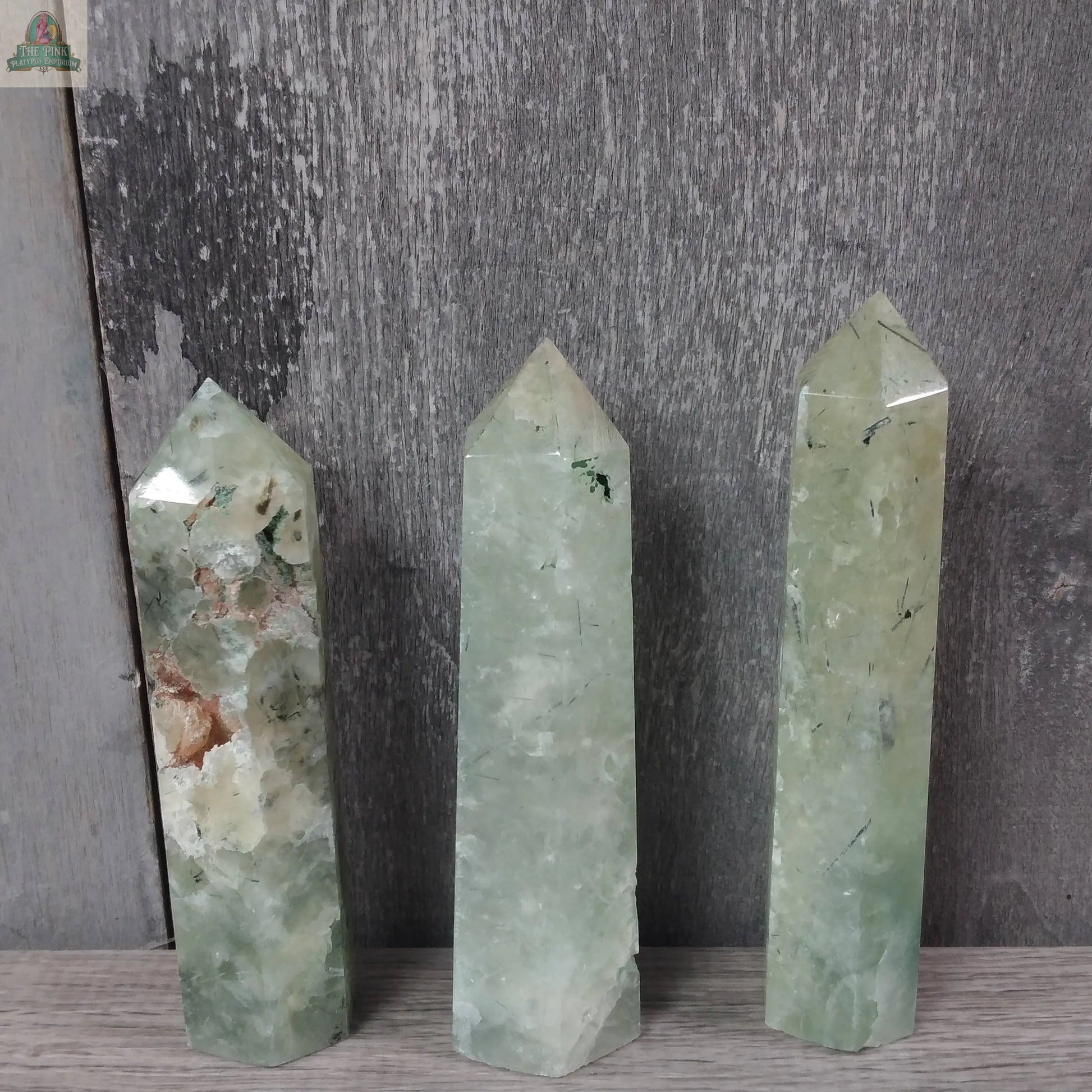 Three Coffee Bean Jasper obelisks from Keystone Crystals stand upright on a gray wooden surface with a textured gray wood background. These bulk 4 to 5 oz gemstone towers are part of the Gemstone Obelisks collection.