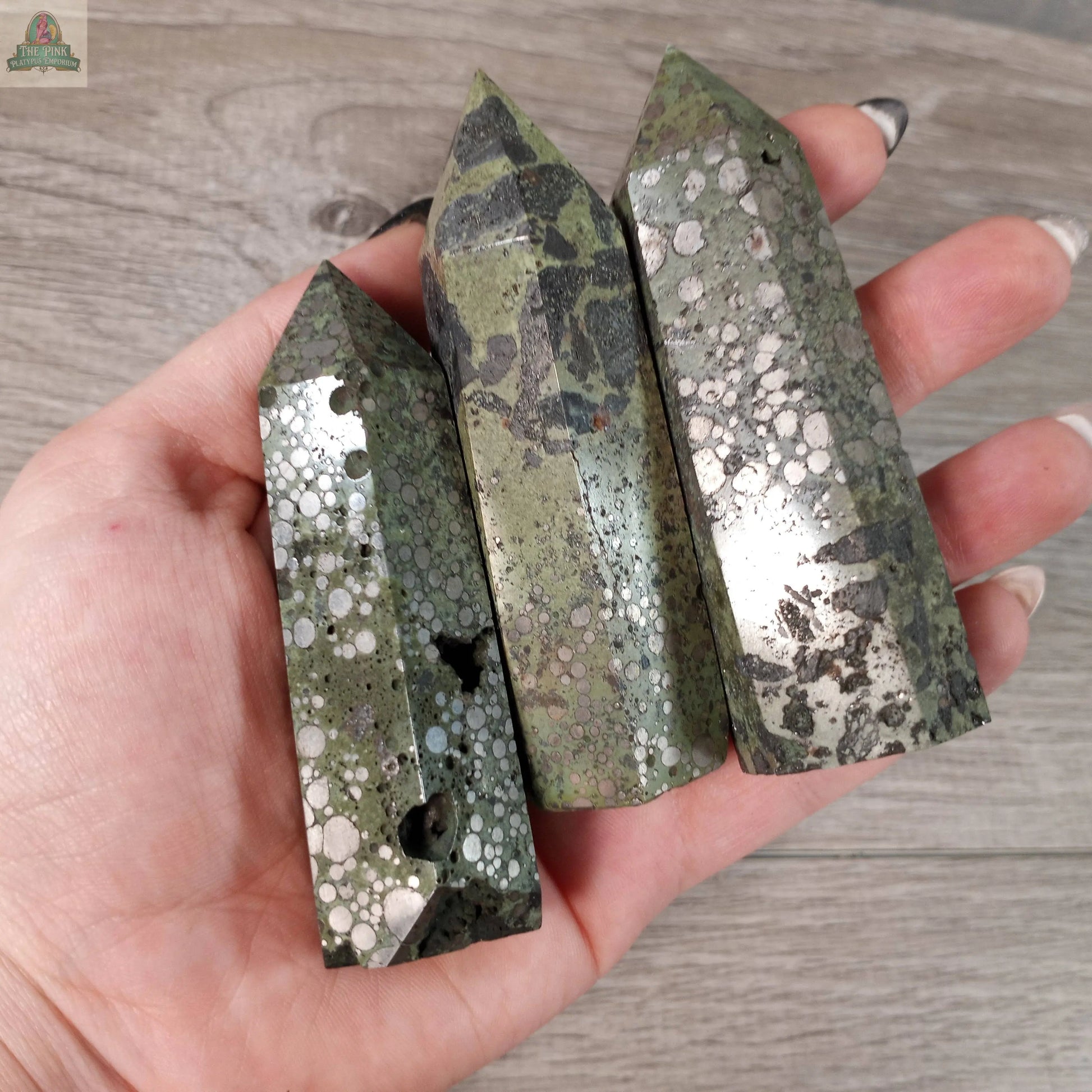 A hand displays three Keystone Crystals Gemstone Obelisks – 4 to 5 oz Bulk Crystal Towers made of green and black Coffee Bean Jasper with circular and spotted patterns, shown against a wooden background.