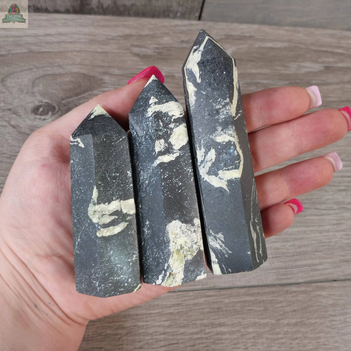 A hand with long, pink nails holds three polished Coffee Bean Jasper crystal towers (4–5 oz) from Keystone Crystals, featuring pointed tips and white veining—perfect gemstone obelisks to elevate any space.