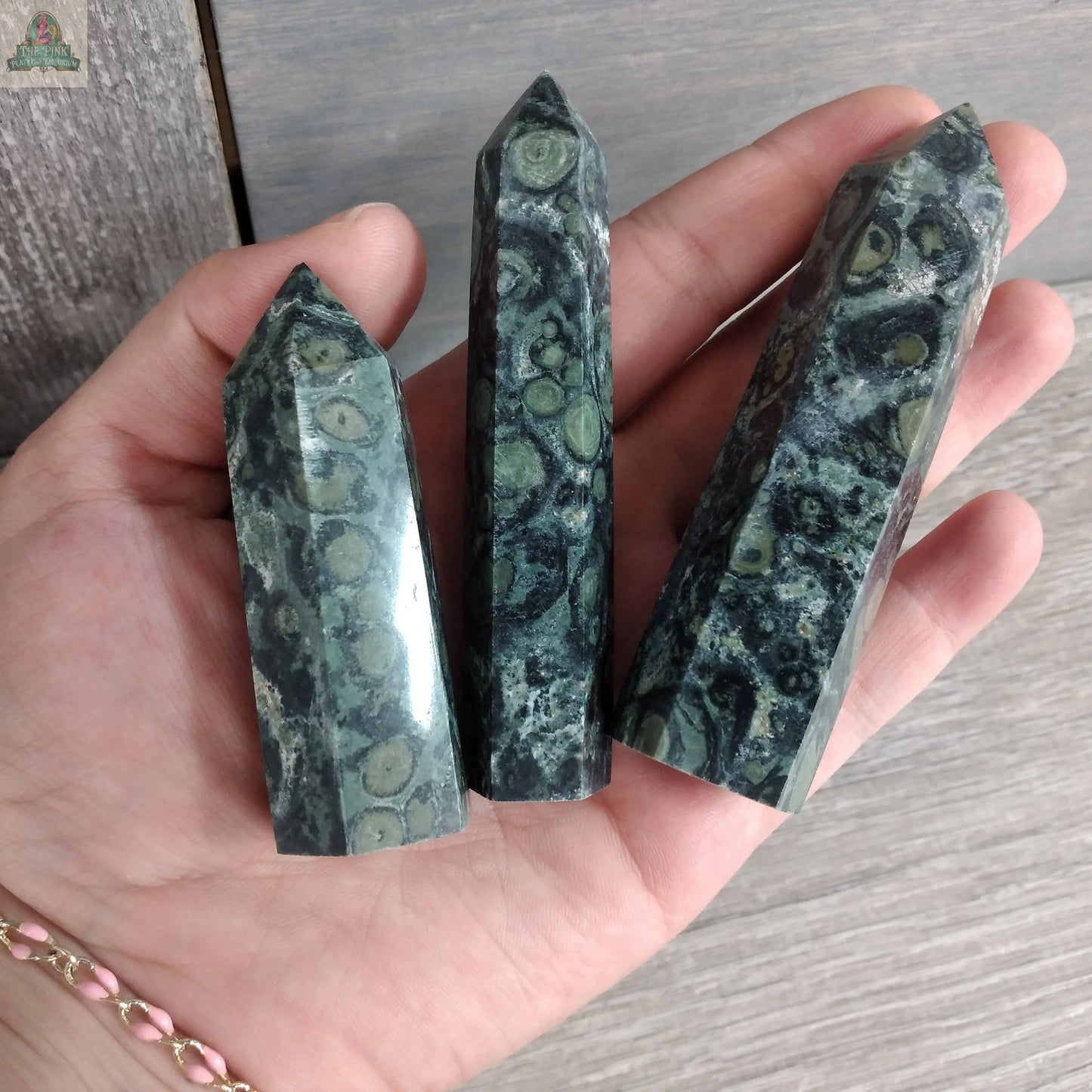 A hand displays three polished Gemstone Obelisks – 4 to 5 oz Bulk Crystal Towers Coffee Bean Jasper by Keystone Crystals, featuring green and black swirling patterns against a wooden background.