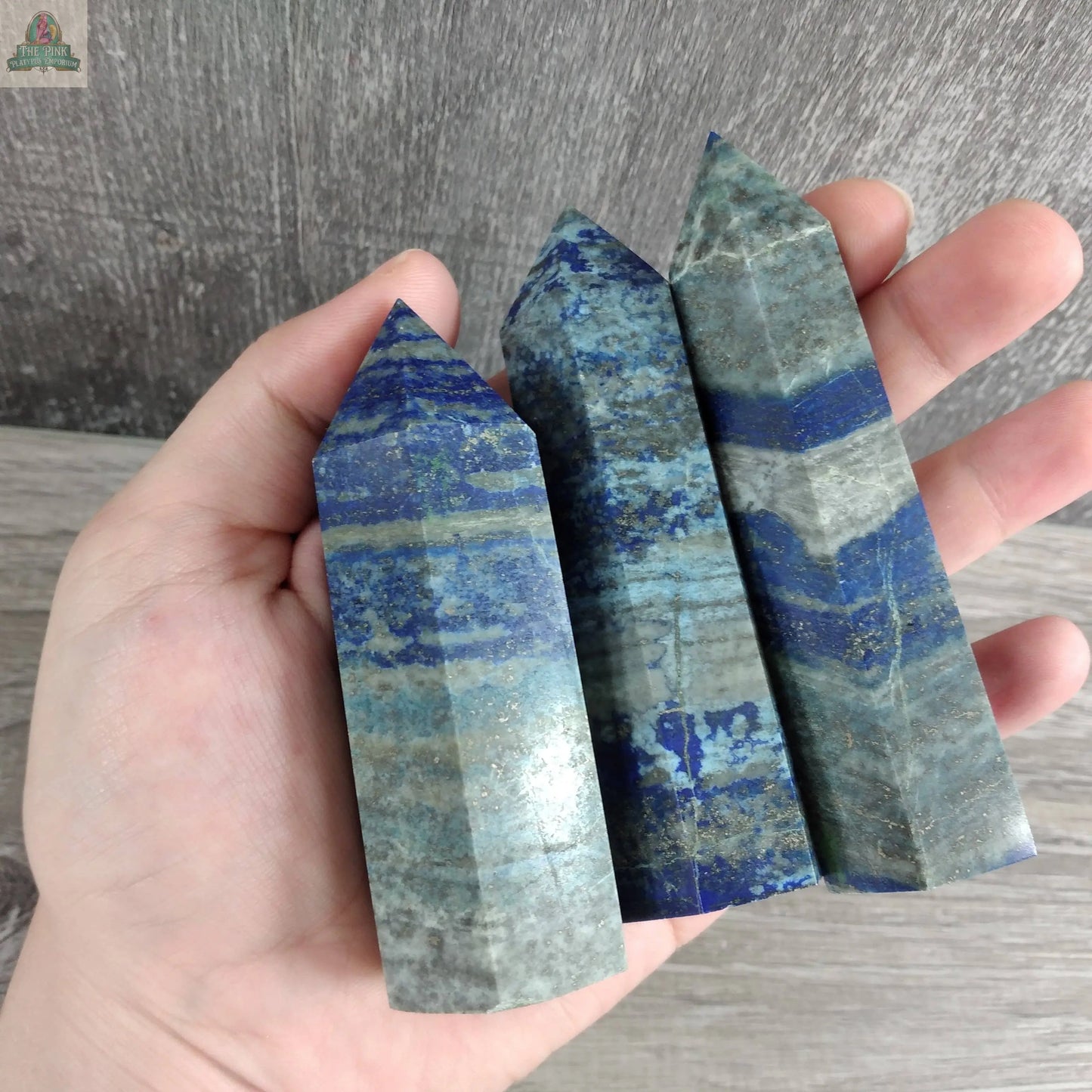 A hand holds three Keystone Crystals Coffee Bean Jasper gemstone obelisks (4–5 oz bulk crystal towers) with blue and gray bands, set against a wooden background.