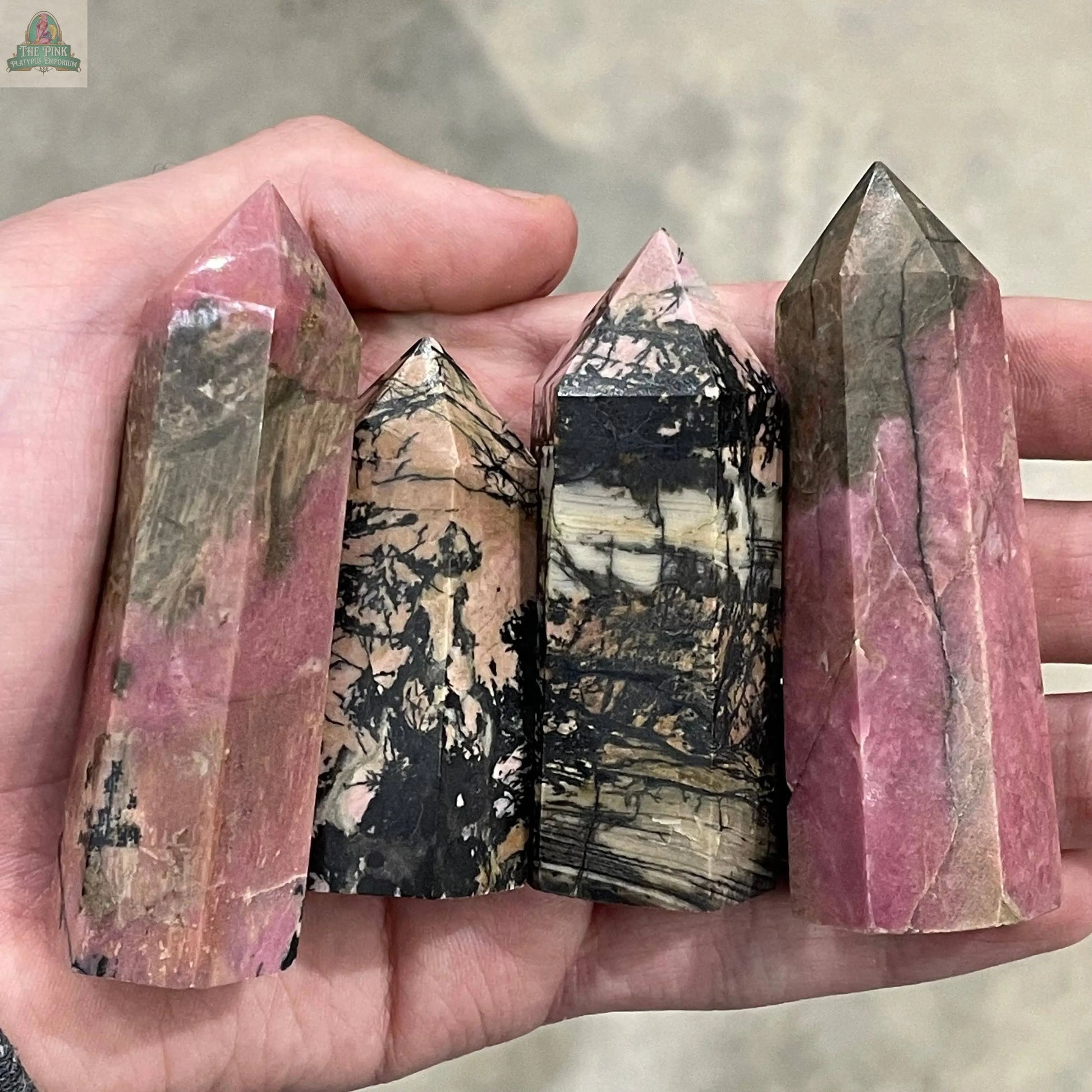 A hand holds four Keystone Crystals Coffee Bean Jasper gemstone obelisks—4 to 5 oz bulk crystal towers with unique patterns—against a neutral background.