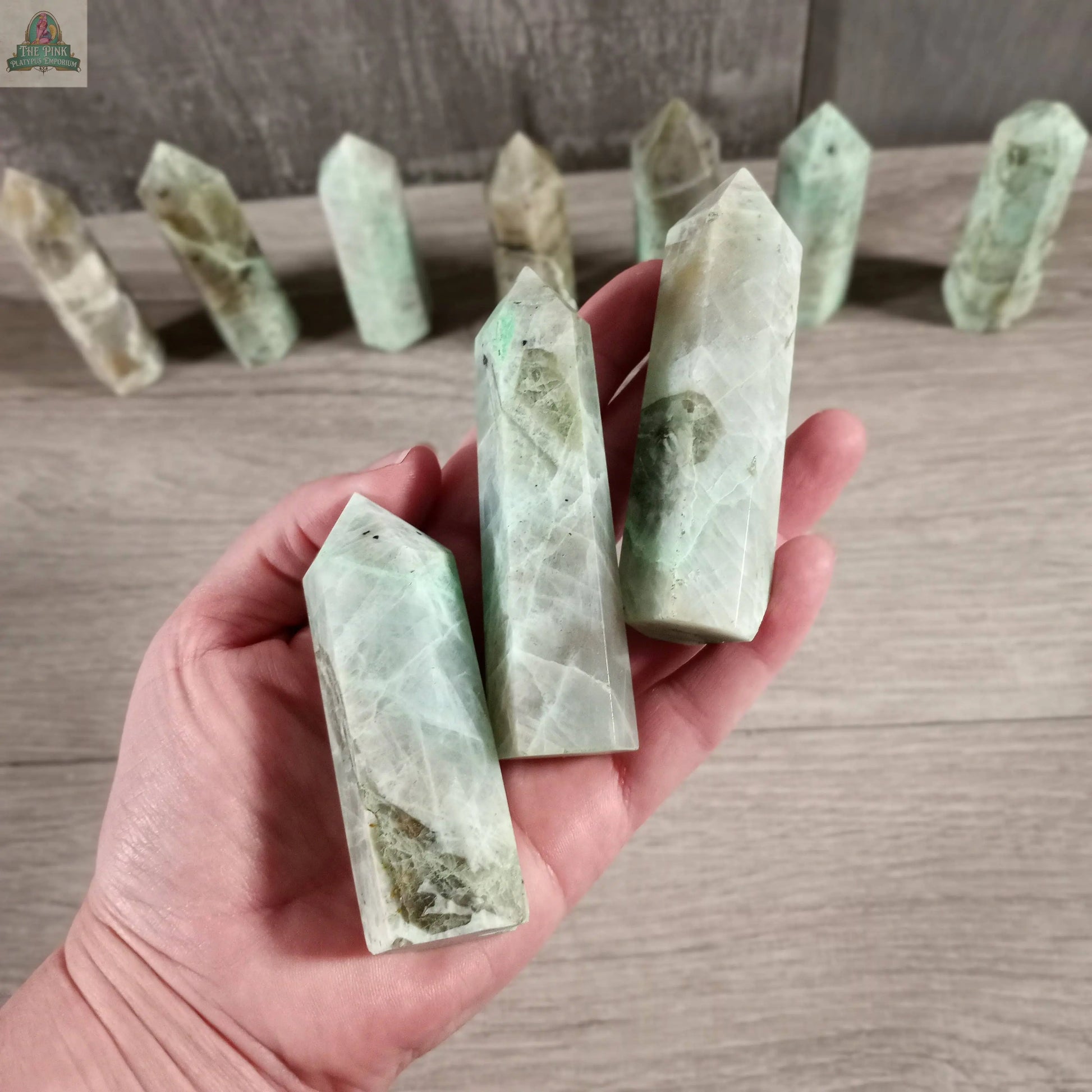 A hand holds three polished Coffee Bean Jasper crystal towers by Keystone Crystals, while several Gemstone Obelisks (4 to 5 oz bulk) are displayed upright on a wooden surface in the background.