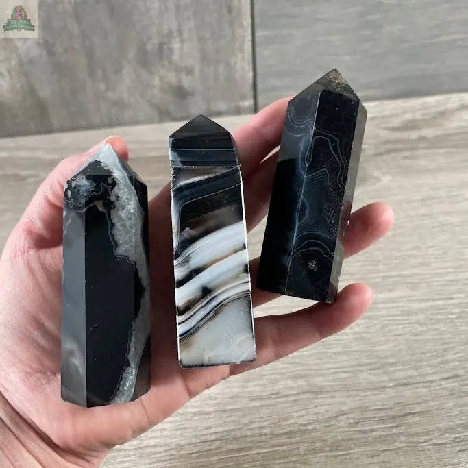 A hand holds three Keystone Crystals Gemstone Obelisks—Coffee Bean Jasper towers (4 to 5 oz each)—showcasing unique black and white banded patterns and pointed tops, displayed against a wooden background.