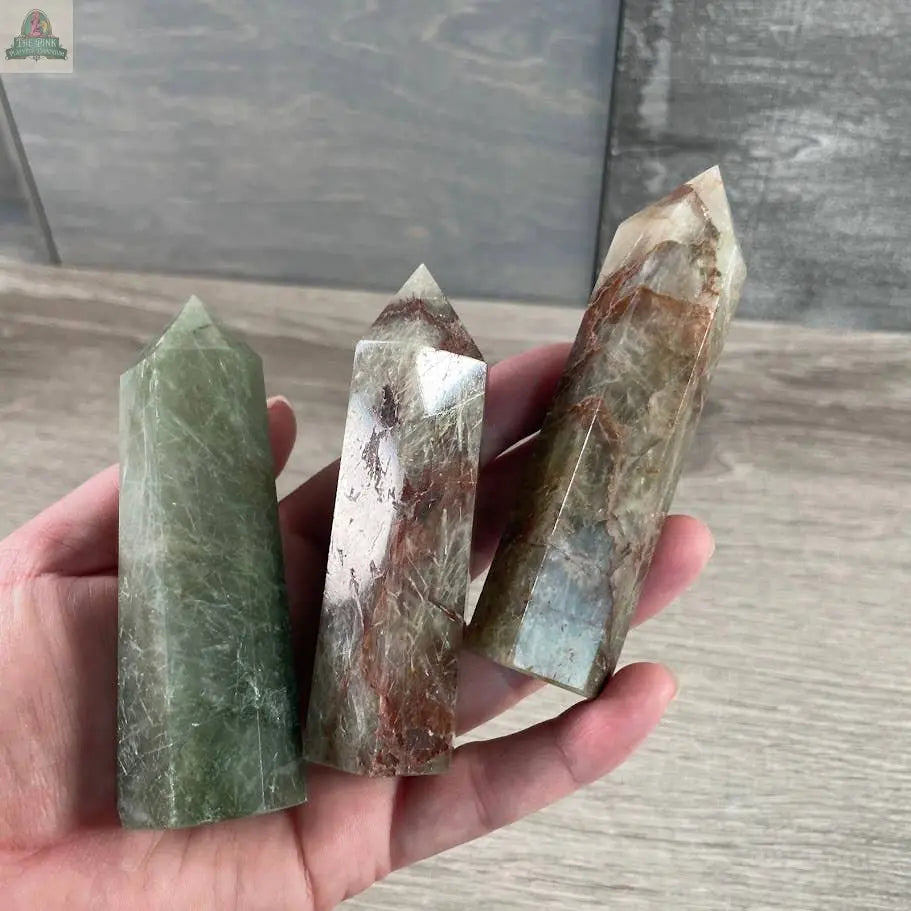 A hand displays three Keystone Crystals Gemstone Obelisks—4 to 5 oz Coffee Bean Jasper bulk crystal towers—featuring natural inclusions in green, brown, and clear shades against a wooden background.