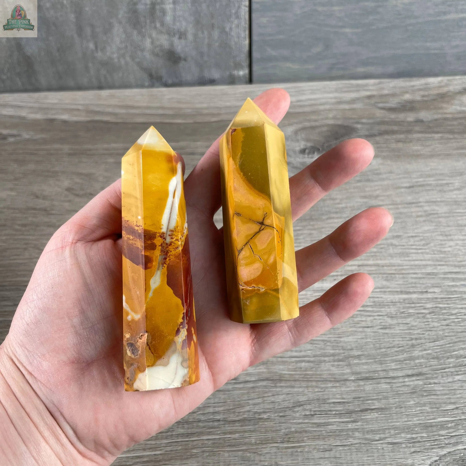 A hand displays two Keystone Crystals Gemstone Obelisks – 4 to 5 oz Coffee Bean Jasper towers with yellow, brown, and white swirls, set against a wooden background.